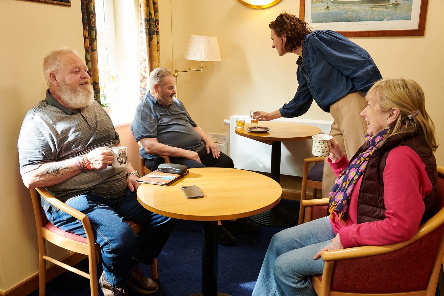 Three elderly people sit around a table, smiling and holding mugs, while a woman stands beside them, serving drinks in a cozy, warmly lit room.