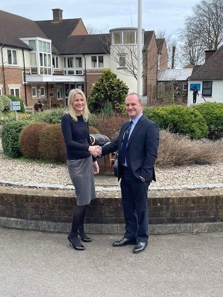 A woman and a man in business attire shake hands and smile outside a building, standing in front of a garden with shrubs and a low brick wall.