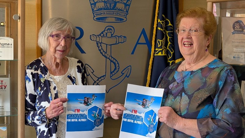 Two older women stand indoors, smiling and holding “I’m Skydiving for Royal Alfred” flyers. Behind them is a wall with a large anchor emblem and flags, suggesting a nautical or military setting.
